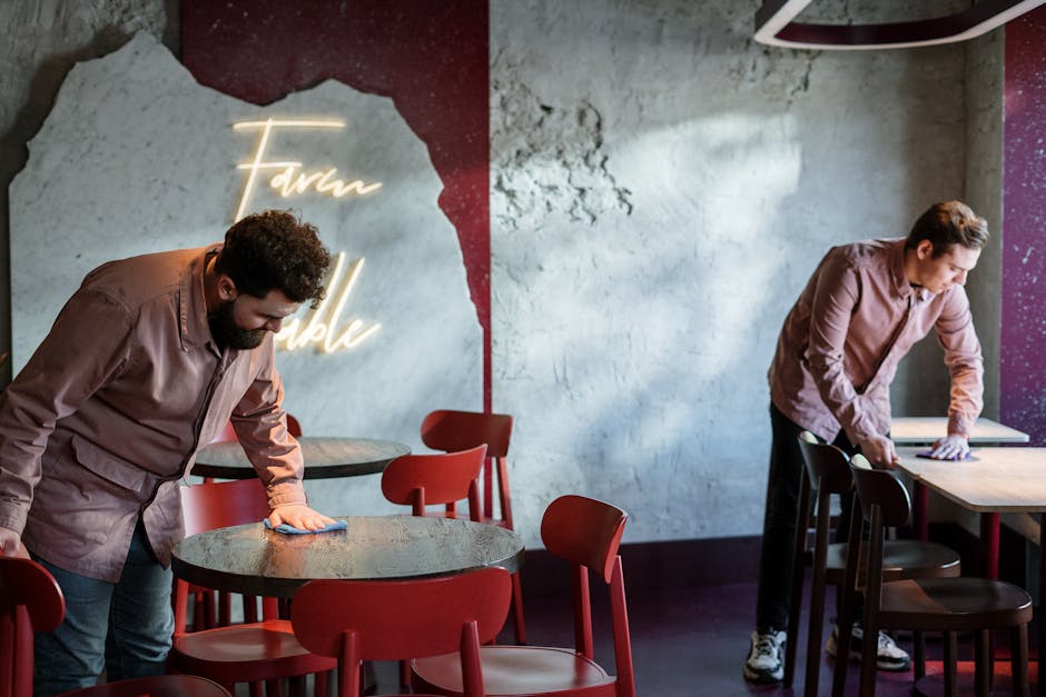 Two staff members cleaning tables in a modern café with neon signage and red chairs.