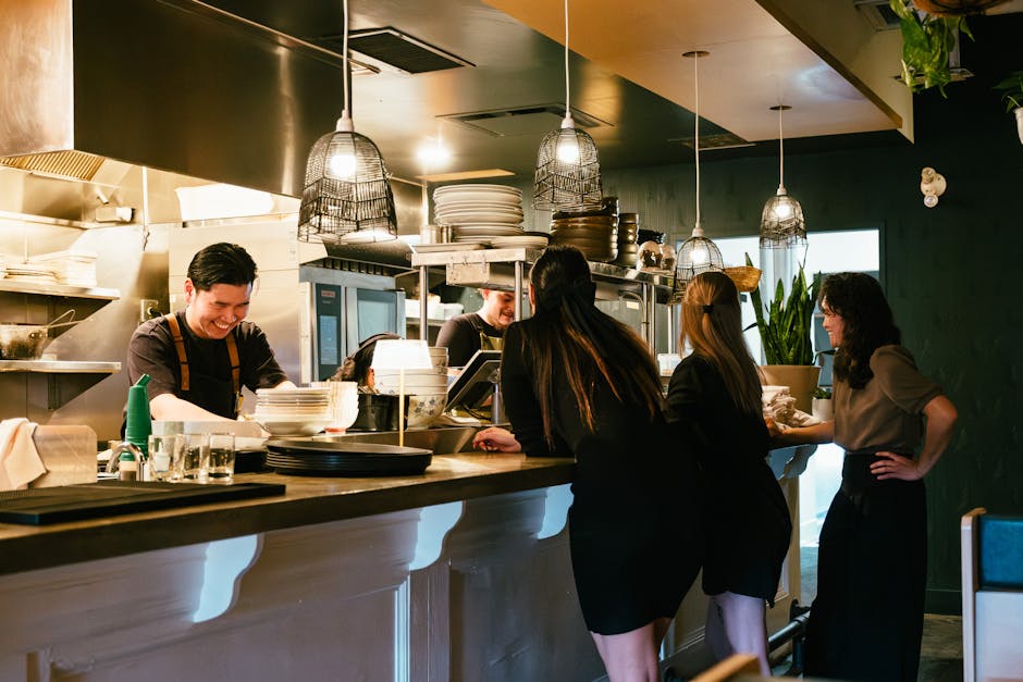 Busy restaurant kitchen with staff preparing meals as patrons watch. Warm lighting and informal dining atmosphere